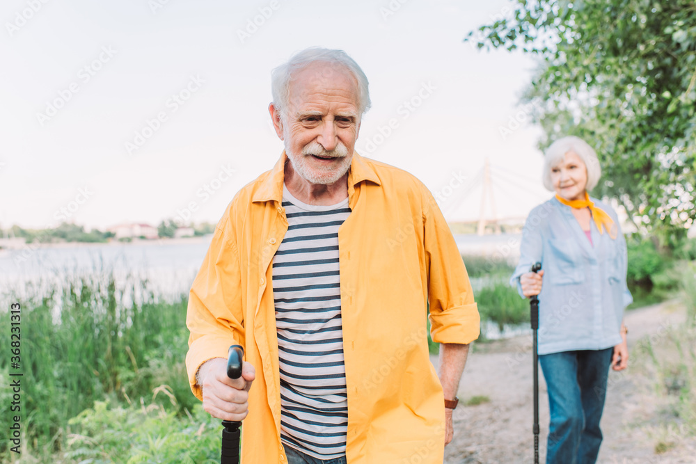 Selective focus of smiling elderly man holding walking stick near wife in park