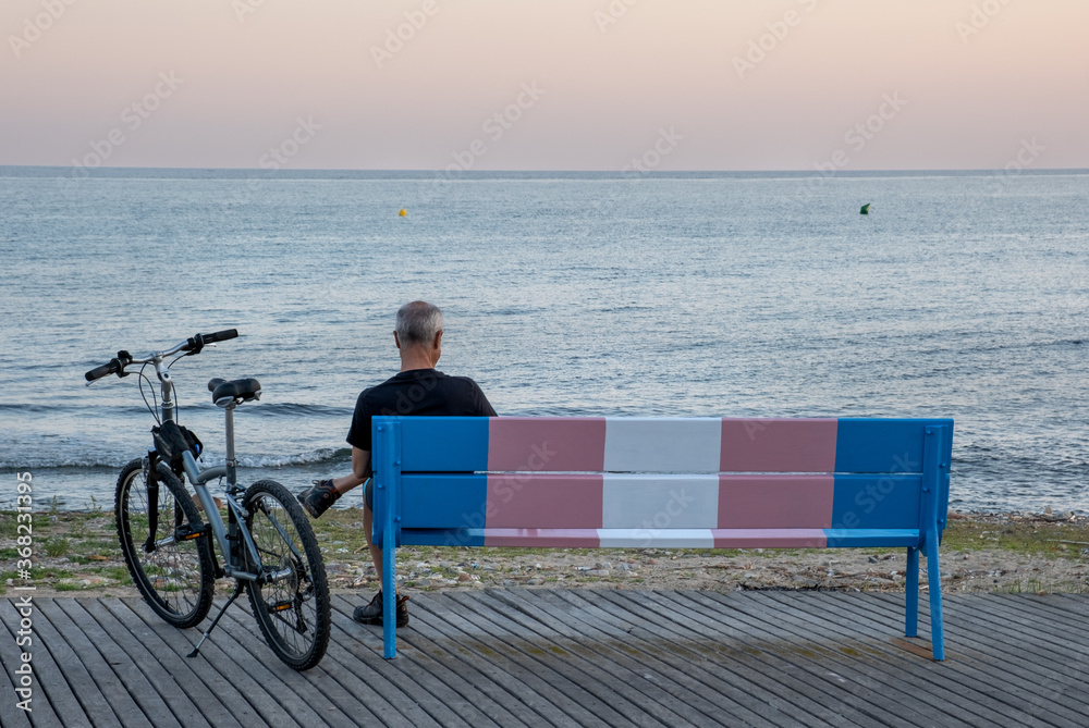 Fototapeta premium Older person with bicycle on a bench painted in the representative colors of transsexuality.