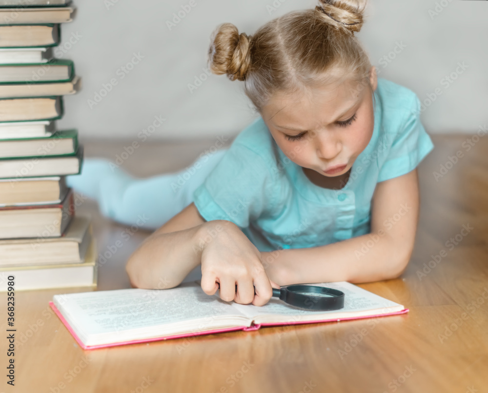 A little blonde schoolgirl is surprised to read book and looks at it through magnifying glass. A pile of books on blurred light background. Back to school. Education. Free time for the child. A little blonde schoolgirl is surprised to read book and looks at it through magnifying glass. A pile of books on blurred light background. Back to school. Education. Free time for the child.