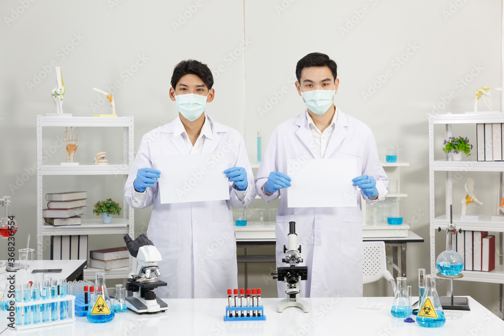 Two Male Scientists wear Face Mask in Lab while holding blank paper. Copy Space. SARS-CoV-2 , Covid-19 THEME. Two Male Scientists wear Face Mask in Lab while holding blank paper. Copy Space. SARS-CoV-2 , Covid-19 THEME.