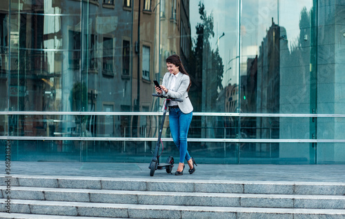 Businesswoman with electric scooter standing in front of modern business building looking at phone. 
