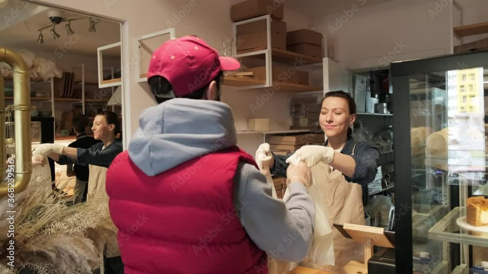 Handheld shot of cheerful young female bakery employee in gloves smiling and handing plastic bags with baked goods to male client in face mask Handheld shot of cheerful young female bakery employee in gloves smiling and handing plastic bags with baked goods to male client in face mask