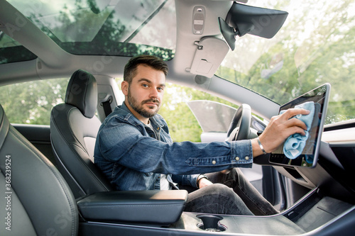Portrait of young smiling handsome Caucasian man with beard sitting inside his modern self-steering car and wiping dust from touchscreen display with microfiber cloth, looking at camera © sofiko14