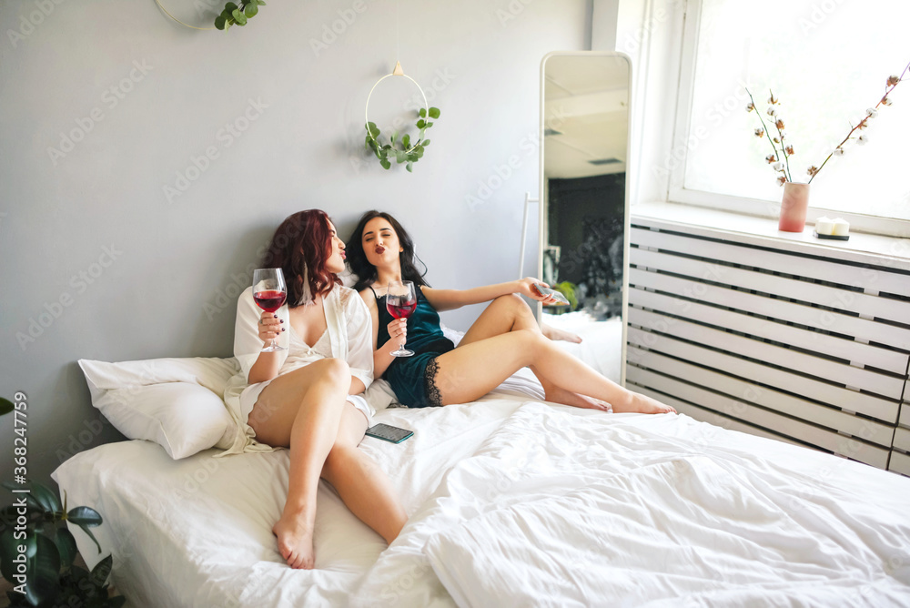 Girlfriends in pajamas are sitting on the bed with wine and telephones. Girlfriends in pajamas are sitting on the bed with wine and telephones.