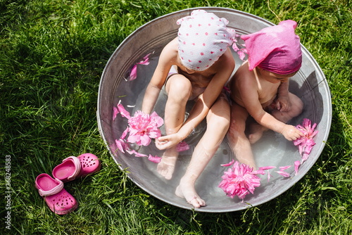 Happy children take a bath in the garden, with peony flowers. Two little sisters bathe with pink flowers in a round basin, outdoors