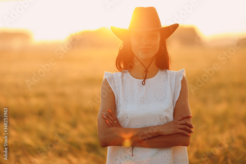 Woman farmer in cowboy hat at agricultural field on sunset with sunflare.