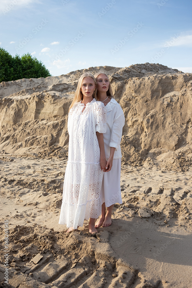 Fototapeta premium two young pretty twins with long blond hair posing at sand quarry in elegant white dress, skirt, jacket. stylish fashion photoshoot, summer photosession. identical sisters spend time together outdoors
