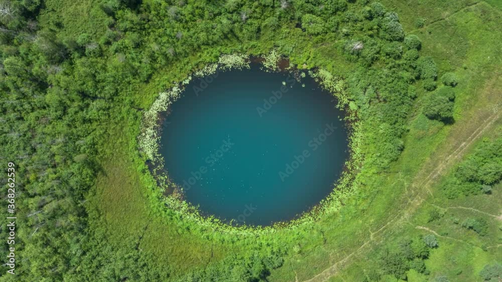 Aerial top down hyperlapse amazing lake of round shape. Cloudy sky reflected in clear turquoise water of pond surrounded by trees and plants. Ripple on water surface, windy sunny summer day