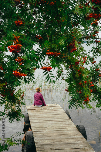 Autumn day by the lake in Karelia. A girl in a pink jacket is sitting under the dense branches on a pier by the lake. photo