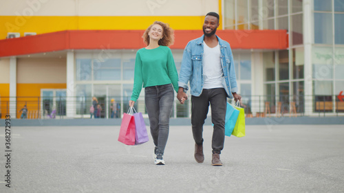 Young diverse couple holding hands when walking outdoors with paper bags after shopping