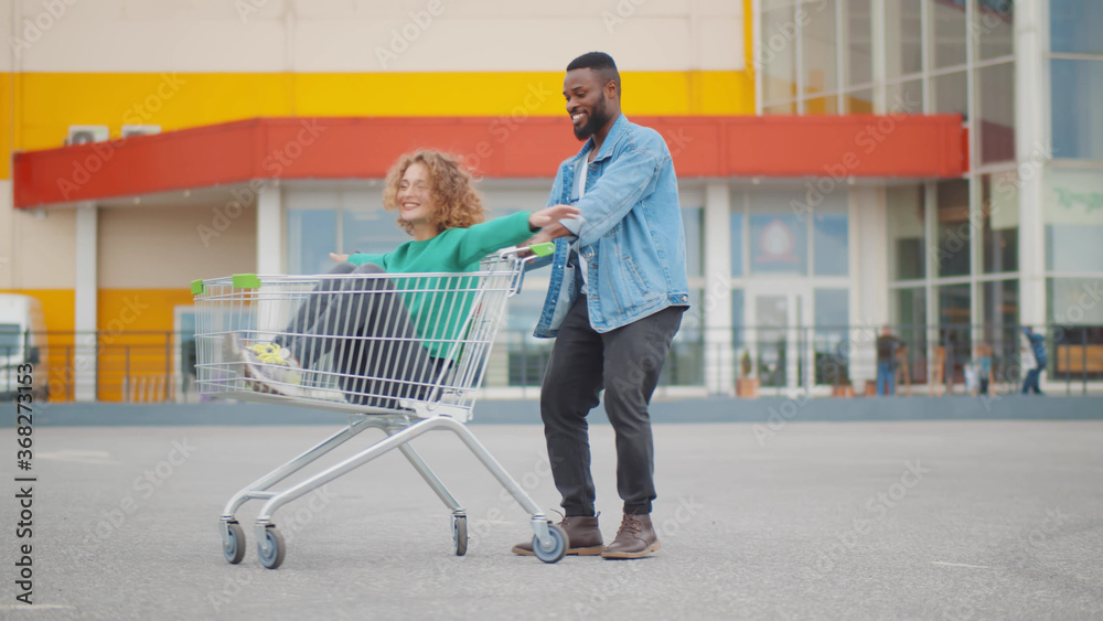 Multiethnic young couple racing with shopping car in parking lot Multiethnic young couple racing with shopping car in parking lot