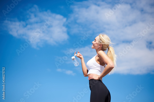 Young blond fit woman, wearing black leggings and white top,drinking water from glass bottle outside with blue sunny sky on background. Female fitness training in summer. Healthy lifestyle concept.