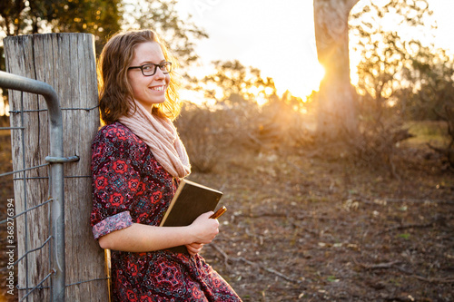 Happy young writer holding pen and notebook leaning on fence post in paddock