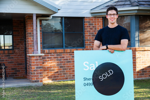 grinning young man with sold sign in front of newly purchased house