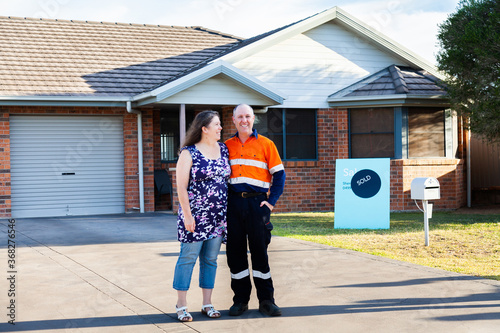 Happy middle aged couple standing infront of house with sold real estate sign photo