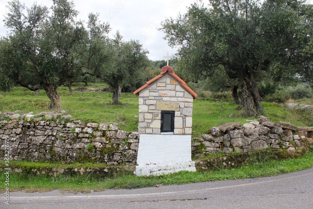 Obraz premium A small country iconostasis, church-like small-scale replica for candle-lighting with olive grove in the background in Messinia region, southwestern Greece.