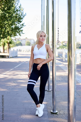 Young fit blond woman, wearing white top and black leggings, leaning on metal pole in city park in summer. Sportswoman, resting relaxing after morning fitness exercise training. Healthy lifestyle.