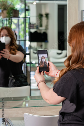 Young hairdresser customer protected by a face mask, taking a selfie of her new look in front of the mirrow. New normality