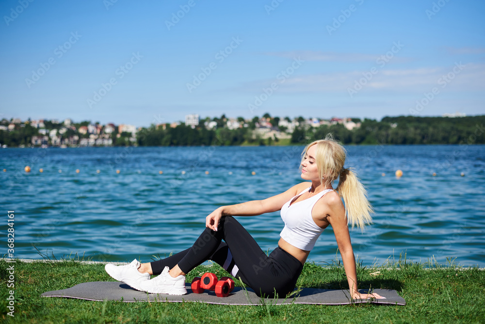 Young blond woman, wearing black leggings and white top, sitting on grey yoga mat by city lake in summer morning, relaxing resting after sport training outside on fresh air. Healthy lifestyle.