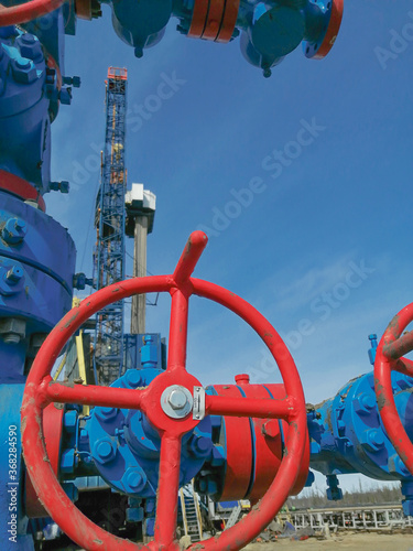 Fountain fittings of a gas well with high-pressure gate valve handwheels. The drilling rig is in the background. Some details of the composition are blurred. Summer day, blue sky.