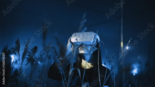 Girl uses VR glasses or headset at modern interactive exhibition in blue neon light outdoor, among wheat field. Future technology, 3D graphics, virtual and augmented reality, concept photo