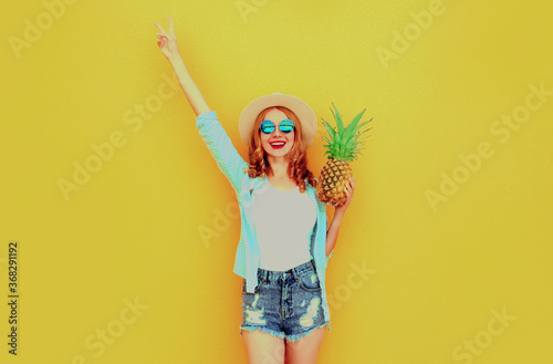 Summer image happy woman raising her hands up with pineapple having fun wearing a straw hat, shorts over colorful yellow background