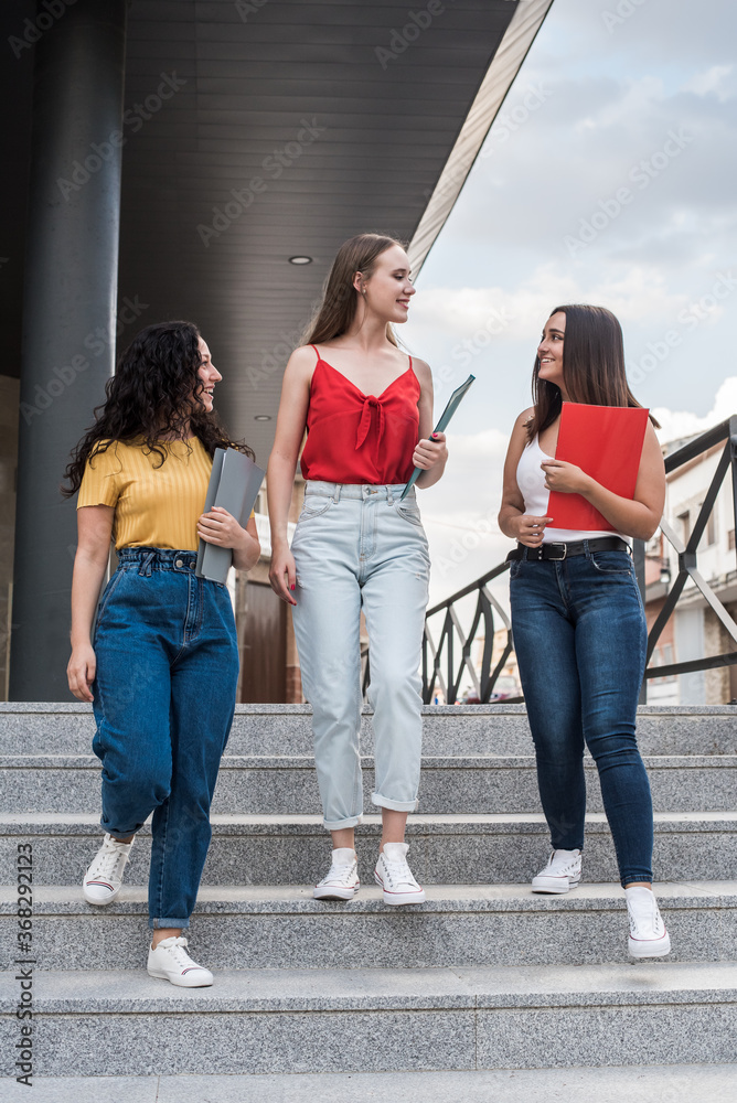 Obraz premium Young women after class talking as they go down campus stairs. Girls dressed in casual clothes with folders in hands