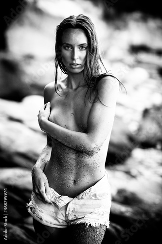 Summertime recreation concept. Beautiful young sexy woman with fit trained slim body wearing blue shorts stands on a beach. Fashion female model poses by the sea. Black and white monochrome.