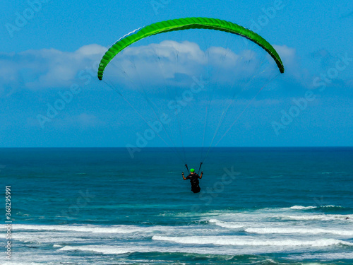 Para glider soars over the beach, waves and hills at Murawai Beach, Auckland, New Zealand