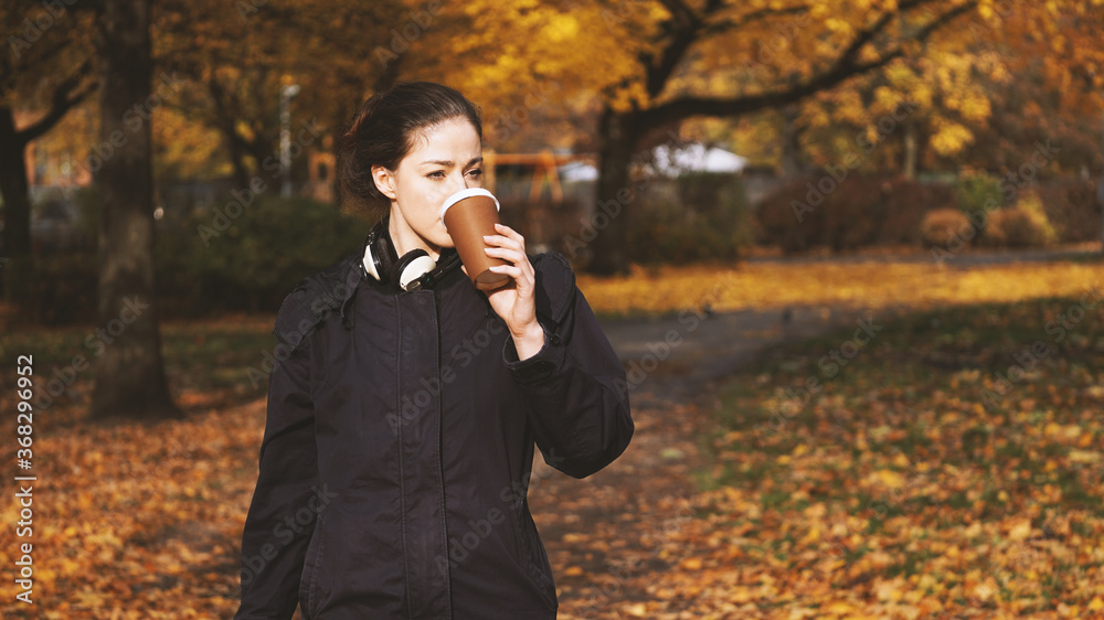 young woman drinking coffee to go from disposable cup while walking in park in autumn young woman drinking coffee to go from disposable cup while walking in park in autumn