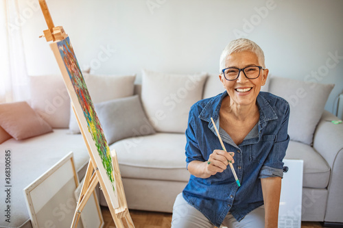 Portrait of attractive middle aged professional female painter with gray hair working at home standing in front of easel. Happy mature woman painting and watching at camera.