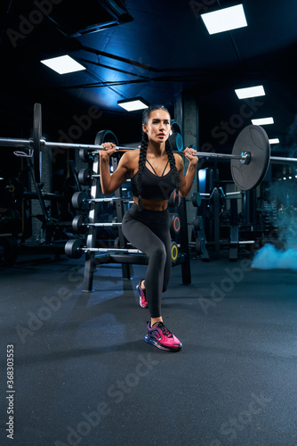 Female bodybuilder doing lunges using barbell in gym.
