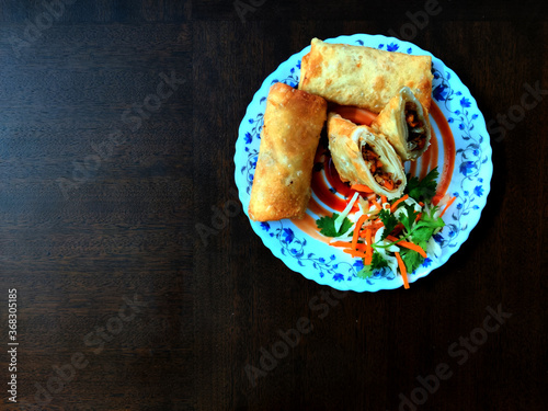 Top view of served Spring Rolls with chilli sauce and fresh chopped vegetables in a white plate on a wooden table