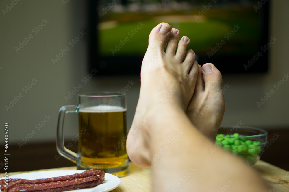 Feet on the table in front of the TV, On the table a mug of beer, a snack in the background of the TV. Rest after work, Friday evening, before weekends, holidays at home alone. Feet on the table in front of the TV, On the table a mug of beer, a snack in the background of the TV. Rest after work, Friday evening, before weekends, holidays at home alone.