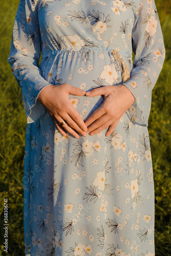 Portrait of young pregnant woman at the field. A woman in a blue dress laid her hands in the form of a heart. Happy mom