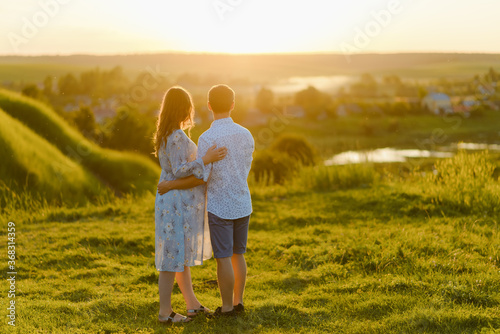A young beautiful pregnant woman in a long dress and her husband standing together in the hill at sunset. Happy family