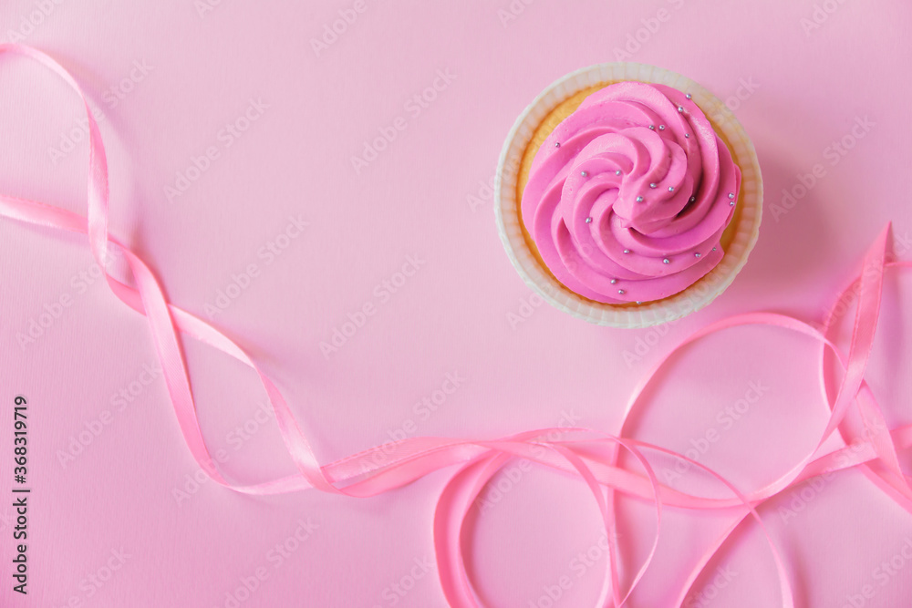 A beautiful cupcake with bright pink cream and a little silver decoration on a pink background. Pink ribbon for decor in the foreground lies in waves. Festive background with free space. A beautiful cupcake with bright pink cream and a little silver decoration on a pink background. Pink ribbon for decor in the foreground lies in waves. Festive background with free space.