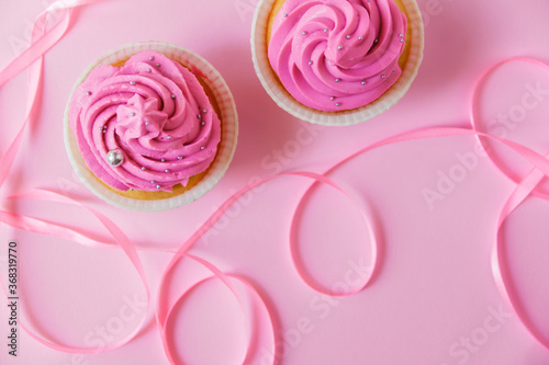 A beautiful cupcakes with bright pink cream and a little silver decoration on a pink background. Pink ribbon for decor in the foreground lies in waves. Festive background with free space.