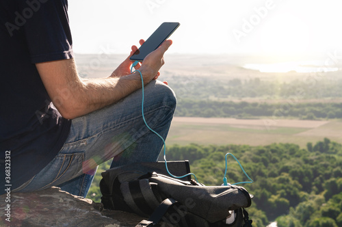 Man on a hike uses smartphone while charging from the power bank on the rock at dawn. Healthy lifestyle and communication.