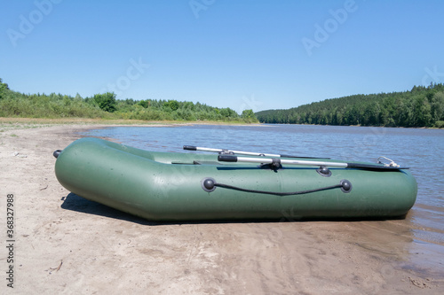 An inflatable rubber boat stands on the banks of a river or lake surrounded by forest. Sunny daytime summer landscape. Background for river walks on water or a concept for fishing. Alone with nature.