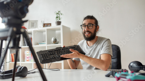 Male technology blogger in glasses looking amazed, holding keyboard while recording video blog or vlog about new gadgets at home