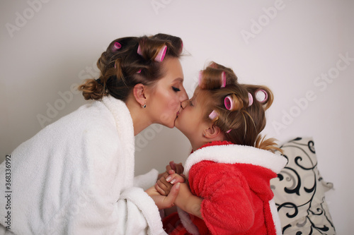 Mom and daughter are sitting in their bathrobes on the bed in their bedroom. They have curlers on their heads.