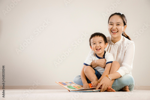 Asian mother and child reading book together concept. Vietnamese mum and son sitting on floor, laughing while flipping pages of a story book, white background
