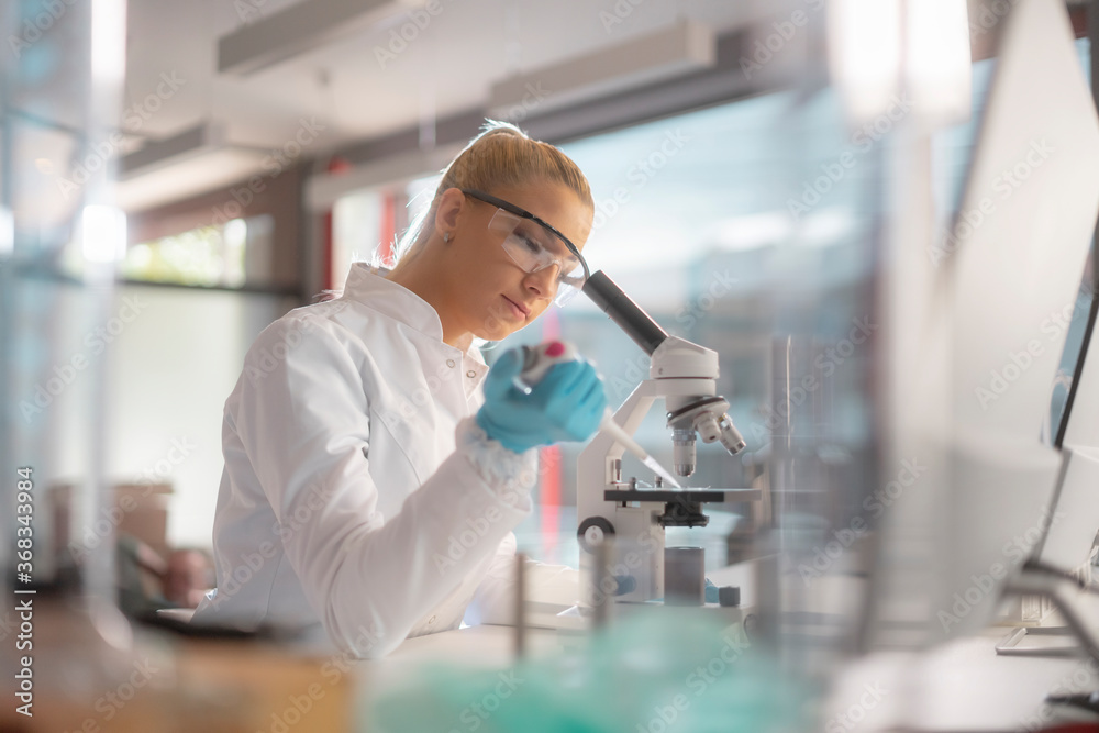 A female scientist wearing a face mask using a pipette in a laboratory studying a sample with a microscope. A female scientist wearing a face mask using a pipette in a laboratory studying a sample with a microscope.