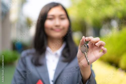Asian real estate agent or realtor woman smiling and holding red file with showing house key in front of thier house in summer day.