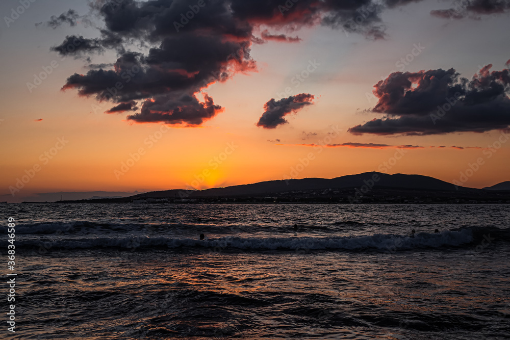A bright sea sunset smoothly leaves the Bay, going behind the mountains of the resort city of Gelendzhik. Colorful summer landscape, beautiful clouds, silhouettes of bathing people covered with waves