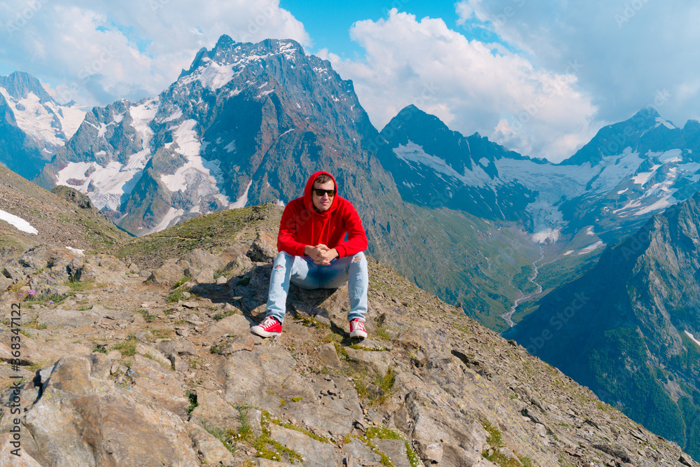 Young man in sunglasses sitting on mountain in sunny weather. Adult male in red hoodie with hood enjoying beautiful view in mountainous area.