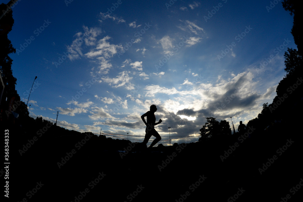 Ein Sportler läuft auf der Laufbahn in einem Stadion und bildet sich als Silhouette vor dem Morgenhimmel ab. Ein Sportler läuft auf der Laufbahn in einem Stadion und bildet sich als Silhouette vor dem Morgenhimmel ab.