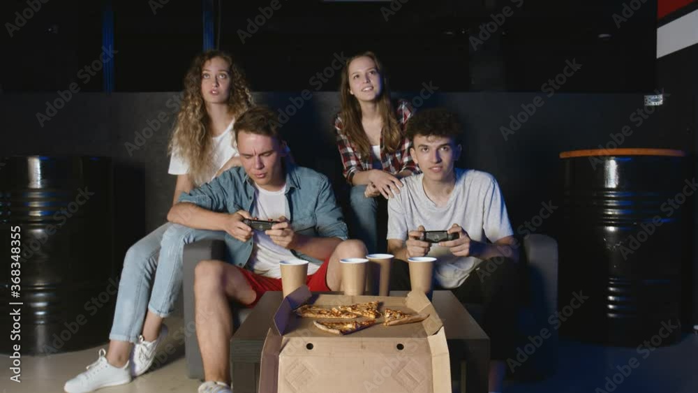 Joyful students girls and guys are having fun with video game at home, one boy is winning laughing and doing high-five, another guy is upset. Joyful students girls and guys are having fun with video game at home, one boy is winning laughing and doing high-five, another guy is upset.