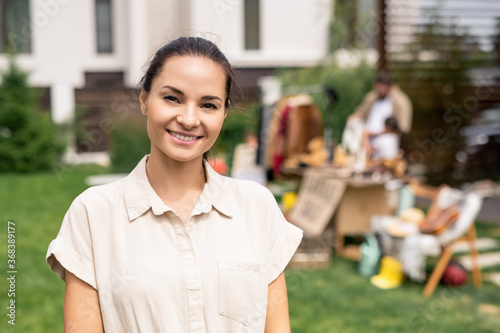 Portrait of smiling attractive young woman in beige blouse visiting garage sale to buy goods for interior